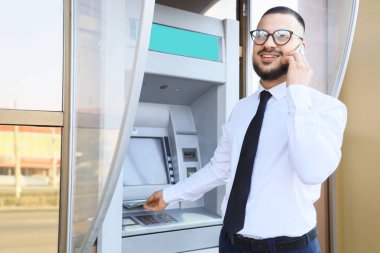 İş adamı dışarıda ATM 'den para çekerken cep telefonuyla konuşuyor.