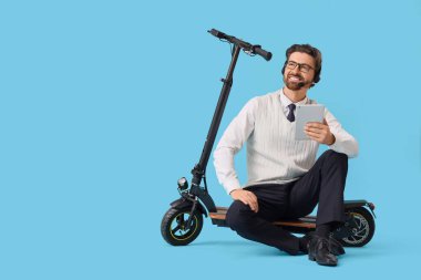 Kulaklıklı, tablet bilgisayarlı yakışıklı bir işadamı mavi arka planda elektrikli scooterın üzerinde oturuyor.