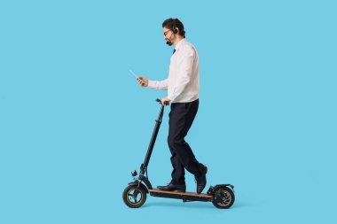 Mavi arka planda elektrikli scooter kullanan, tablet bilgisayarlı yakışıklı bir iş adamı.