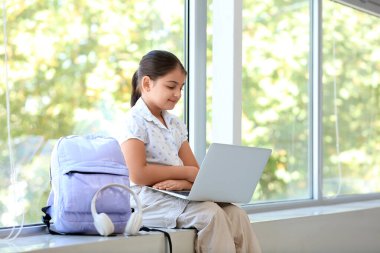 Küçük liseli kız evdeki pencere eşiğinde dizüstü bilgisayarla ders çalışıyor.