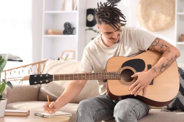 Rastalı genç adam evdeki kanepede gitar çalıyor.