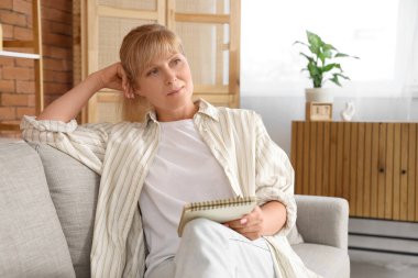 Düşünceli olgun bir kadın evdeki kanepede deftere yazıyor.