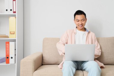 Ofisteki kanepede dizüstü bilgisayarla çalışan genç Asyalı adam.