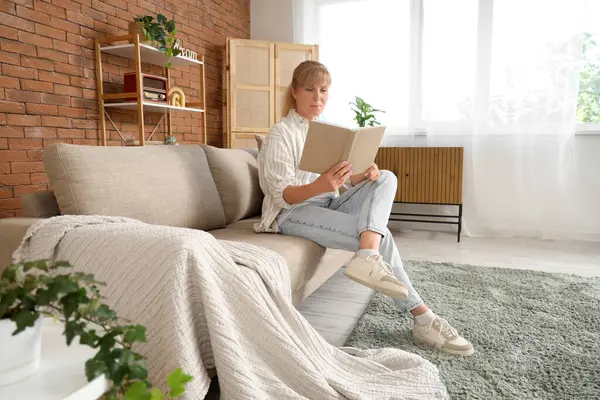 Olgun kadın evdeki kanepede kitap okuyor.
