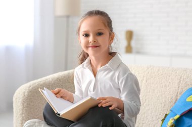 Kitap ve sırt çantasıyla kanepede oturan ve evde ders çalışan sevimli küçük kız.