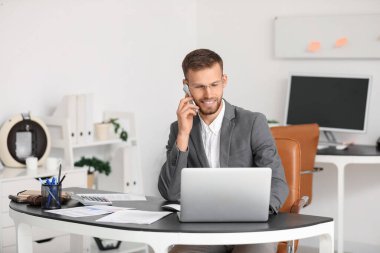 Gözlüklü genç işadamı cep telefonuyla konuşuyor ve iş yerinde belgeler ve bilgisayarla ofiste oturuyor.