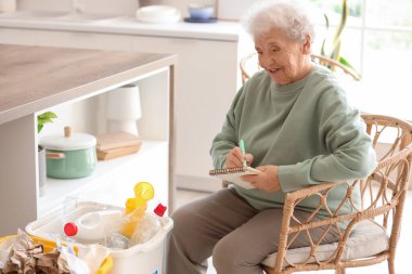 Mutfaktaki not defterine çöp bidonları yazan son sınıf öğrencisi kadın. Atık sıralama kavramı