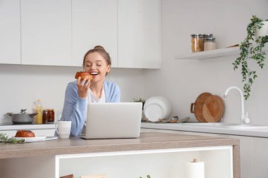 Modern dizüstü bilgisayarlı genç bir kadın mutfakta çörek yiyor.