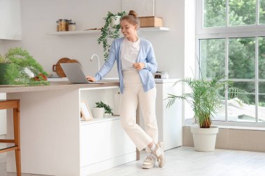 Genç kadın mutfaktaki masada modern dizüstü bilgisayar kullanıyor.