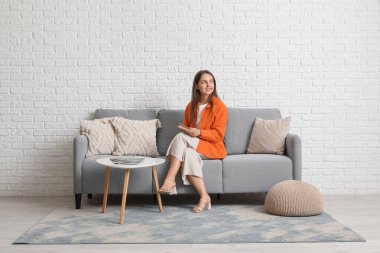 Genç, güzel bir kadın kitap okuyor ve evdeki gri kanepede dinleniyor.