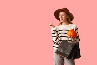Güz kıyafetleri içinde, balkabağı ve alışveriş torbalarıyla pembe arka plandaki bir şeyi gösteren genç bir kadın.