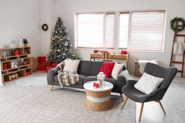 Oturma odasının içi gri kanepe, koltuk, Noel ağacı ve şenlikli süslemelerle dolu.