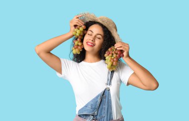 Hasır şapkalı, mavi arka planda olgun üzümlü güzel Afro-Amerikalı kadın.