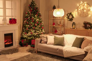 Şenlikli oturma odasının içinde şömine, Noel ağacı ve akşamları parlayan ışıklar.