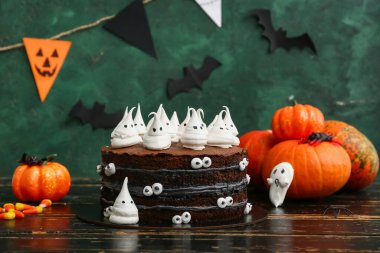 Tatlı Cadılar Bayramı pastası, yeşil arka planda siyah ahşap masada hayaletler, şekerler ve balkabaklarıyla süslenmiş.