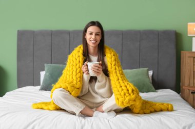 Yatak odasında, sıcak sarı ekoseli ve çay fincanı olan genç ve güzel bir kadın.