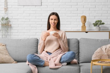 Evdeki kanepede dinlenen pembe ekoseli ve bir fincan kahveli genç kadın.