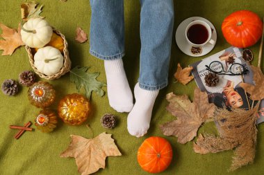 Sonbahar yapraklı sıcak çoraplar içinde kadın bacakları, arka plan olarak yeşil ekose üzerinde bir fincan çay ve balkabağı.