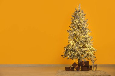 Parlayan ışıklarla süslenmiş Noel ağacı ve sarı duvarın yanında hediye kutuları.