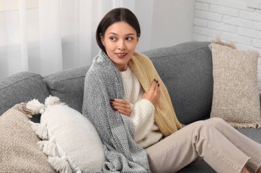 Evdeki kanepede battaniyeli beyaz örülü süveter giyen güzel genç bir kadın oturuyor.