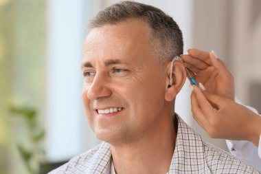 Kadın doktor klinikte adamın kulağına işitme cihazı takıyor.