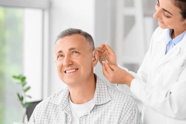 Kadın doktor klinikte adamın kulağına işitme cihazı takıyor.