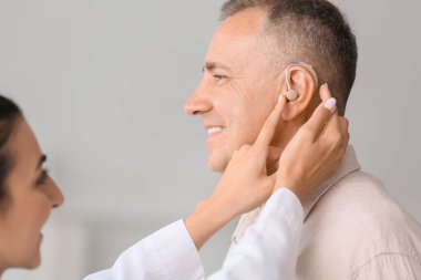 Kadın doktor klinikte adamın kulağına işitme cihazı takıyor.