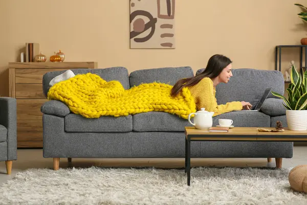 Oturma odasındaki kanepede yatarken dizüstü bilgisayar kullanan, ılık sarı ekoseli güzel genç bir kadın.