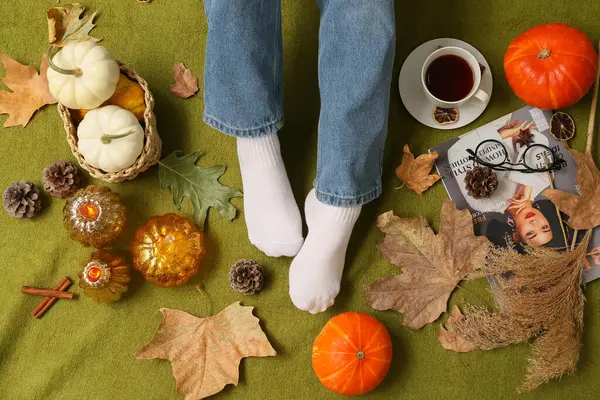Sonbahar yapraklı sıcak çoraplar içinde kadın bacakları, arka plan olarak yeşil ekose üzerinde bir fincan çay ve balkabağı.