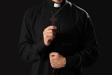 Karanlık arka planda haç ve kutsal kitaba sahip genç rahip, yakın plan.