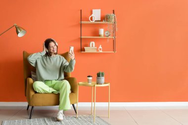 Kulaklığı ve cep telefonu olan genç bir kadın evdeki koltukta oturuyor.