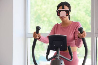 VR gözlüklü genç Afro-Amerikalı kadın spor salonunda egzersiz bisikletinde