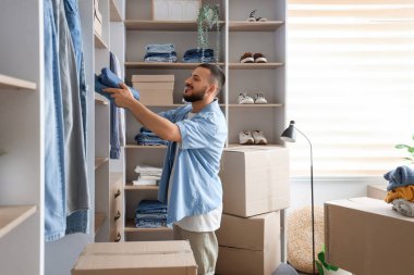 Genç adam gardıroptaki raftan kot pantolon alıyor.