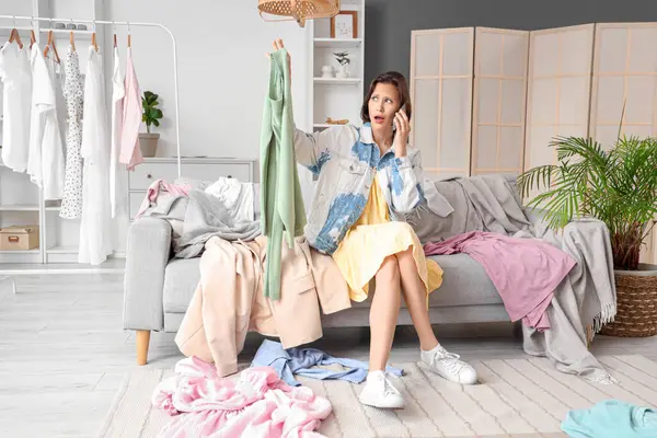 Şok olmuş genç bir kadın elinde kıyafetlerle evdeki koltukta cep telefonuyla konuşuyor.