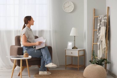 Kitaplı genç bir kadın oturma odasında duvar saatine bakıyor.