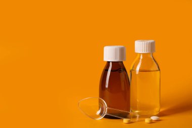 Öksürük şurubu şişeleri, ölçüm kaşığı ve turuncu arka planda haplar.