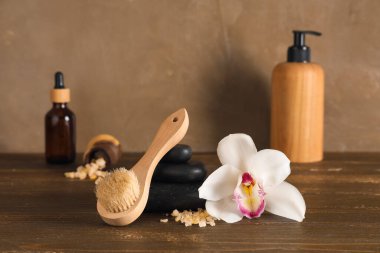 Masaj fırçası, spa taşları, deniz tuzu, orkide çiçeği ve kozmetik ürünleri koyu ahşap masa üzerinde.