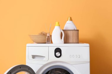 Turuncu duvarın yanında deterjan şişeleri olan modern çamaşır makinesi.