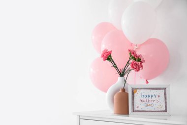 Karanfil çiçekli vazolar, balonlar ve Anneler Günü kartı ışık duvarının yanında raflarda.
