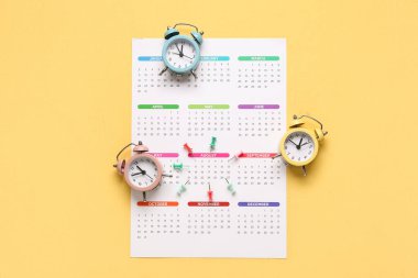 Sarı zemin üzerinde alarm saatleri ve raptiyeler olan bir takvim. Planlama kavramı
