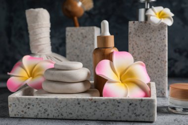 Gri grunge arka planında tepsi, taş, plumeria çiçekleri ve bir şişe kozmetik ürünüyle güzel bir spa kompozisyonu, yakın plan.