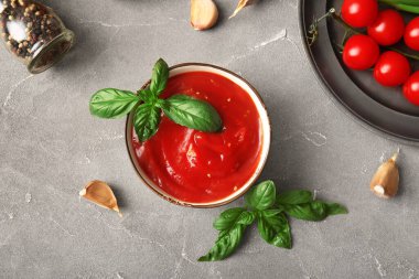Bir kase lezzetli domates sosu, fesleğen yaprağı ve gri arka planda sarımsak.