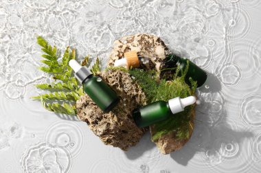 Kozmetik şişeleri, yeşil yapraklar ve beyaz arka planda suda taşlar.