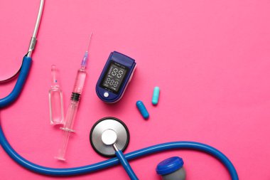 Nabız oksimetresi, steteskop, ampul, haplar ve pembe arka planda şırınga.
