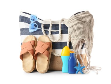 Bikini, güneş gözlüğü, terlik ve beyaz arka planda güneş kremi şişeleriyle çizgili plaj çantası.