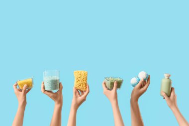 Banyo bombası, sünger, deniz tuzu ve mavi arka planda bir şişe kozmetik ürünü taşıyan kadın elleri.