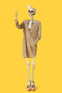 Parti şapkalı ve elbiseli insan iskeleti sarı arka planda el kaldırıyor.