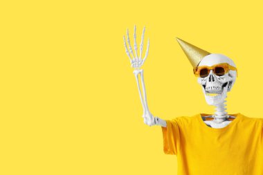 Parti şapkalı ve tişörtlü insan iskeleti sarı arka planda el kaldırıyor.