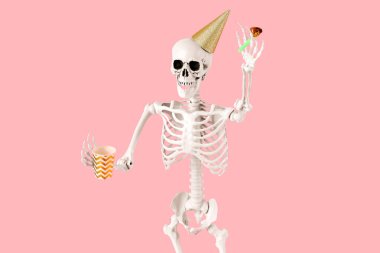 Parti şapkalı insan iskeleti elinde kağıt bardak ve pembe arka planda parti düdüğü, yakın plan.