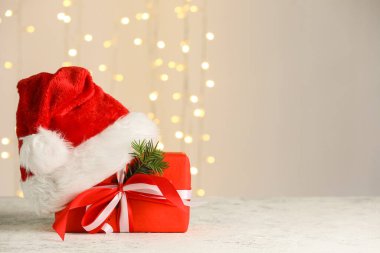 Hediye kutulu kompozisyon, Noel Baba şapkası ve köknar dalı bej duvarın yanındaki gri masanın üzerinde parlayan ışıklar, yakın plan.